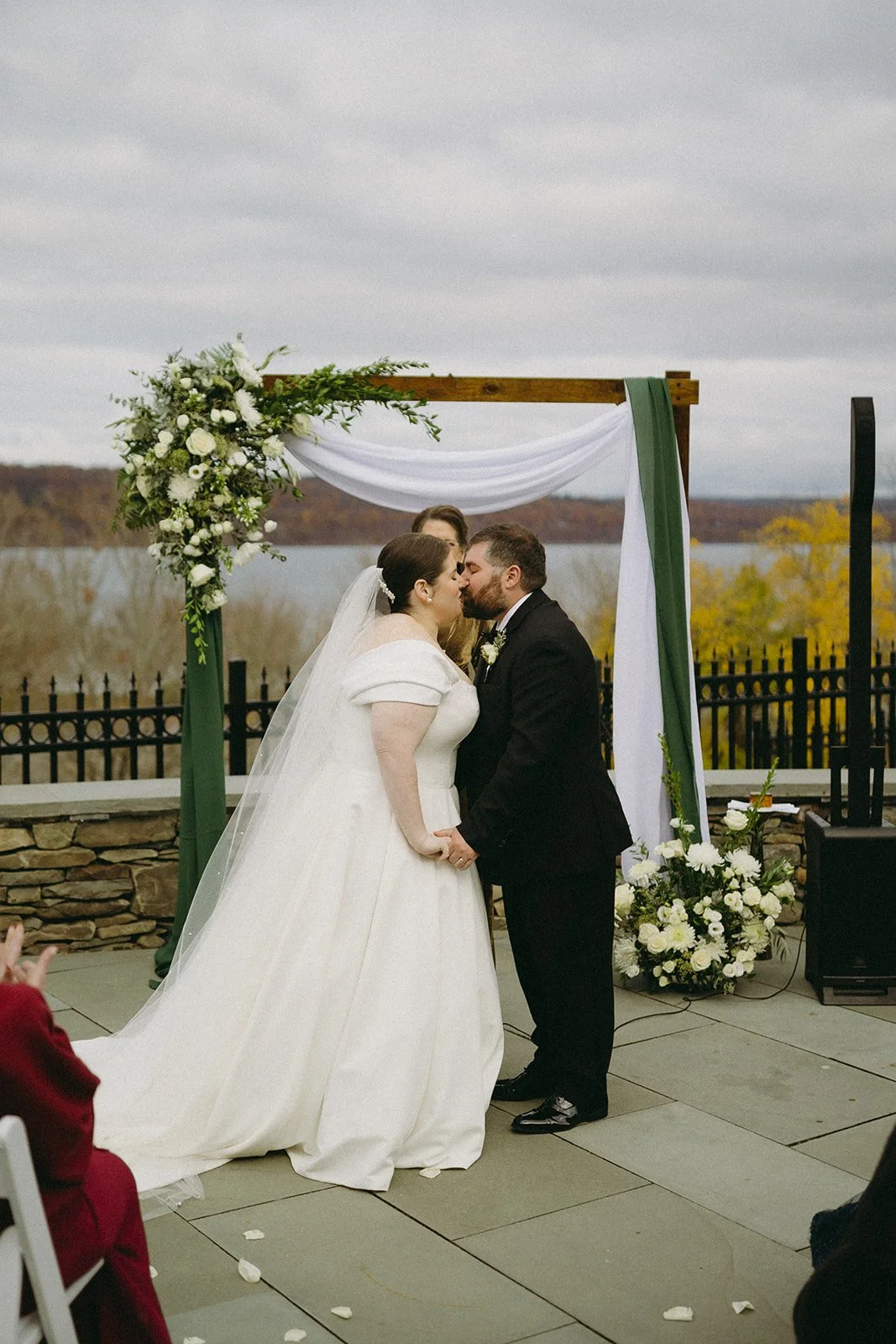 A bride and groom stand before an officiant under a floral arch during an outdoor wedding ceremony, with seated guests in the foreground for an Inn at Taughannock Falls Wedding