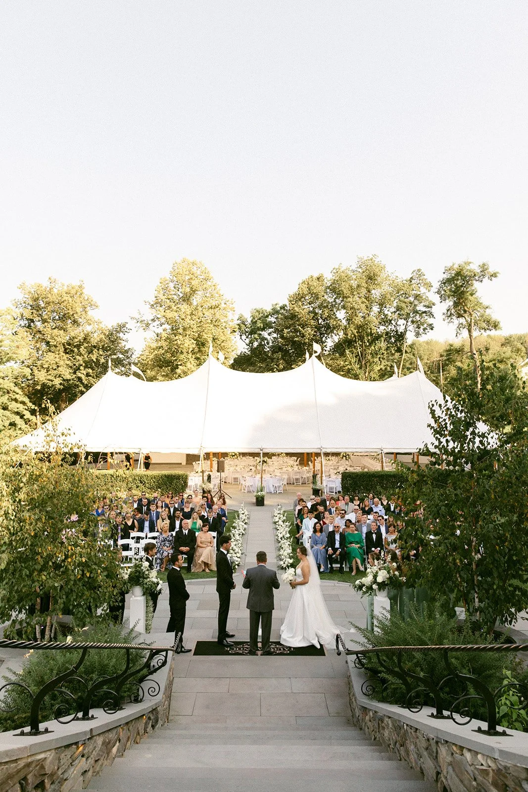 An outdoor wedding ceremony is taking place in front of a large white tent, with guests seated and a couple standing at the altar for an Inn at Taughannock Falls Wedding