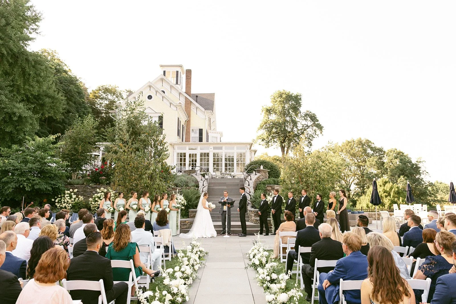 An outdoor wedding ceremony is taking place in front of a large white tent, with guests seated and a couple standing at the altar for an Inn at Taughannock Falls Wedding