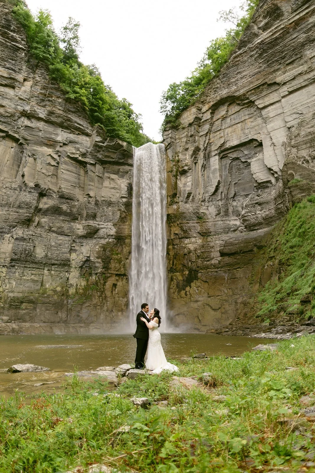 A bride and groom stand together near the base of a tall waterfall, surrounded by rocky cliffs and greenery at Taughannock Falls