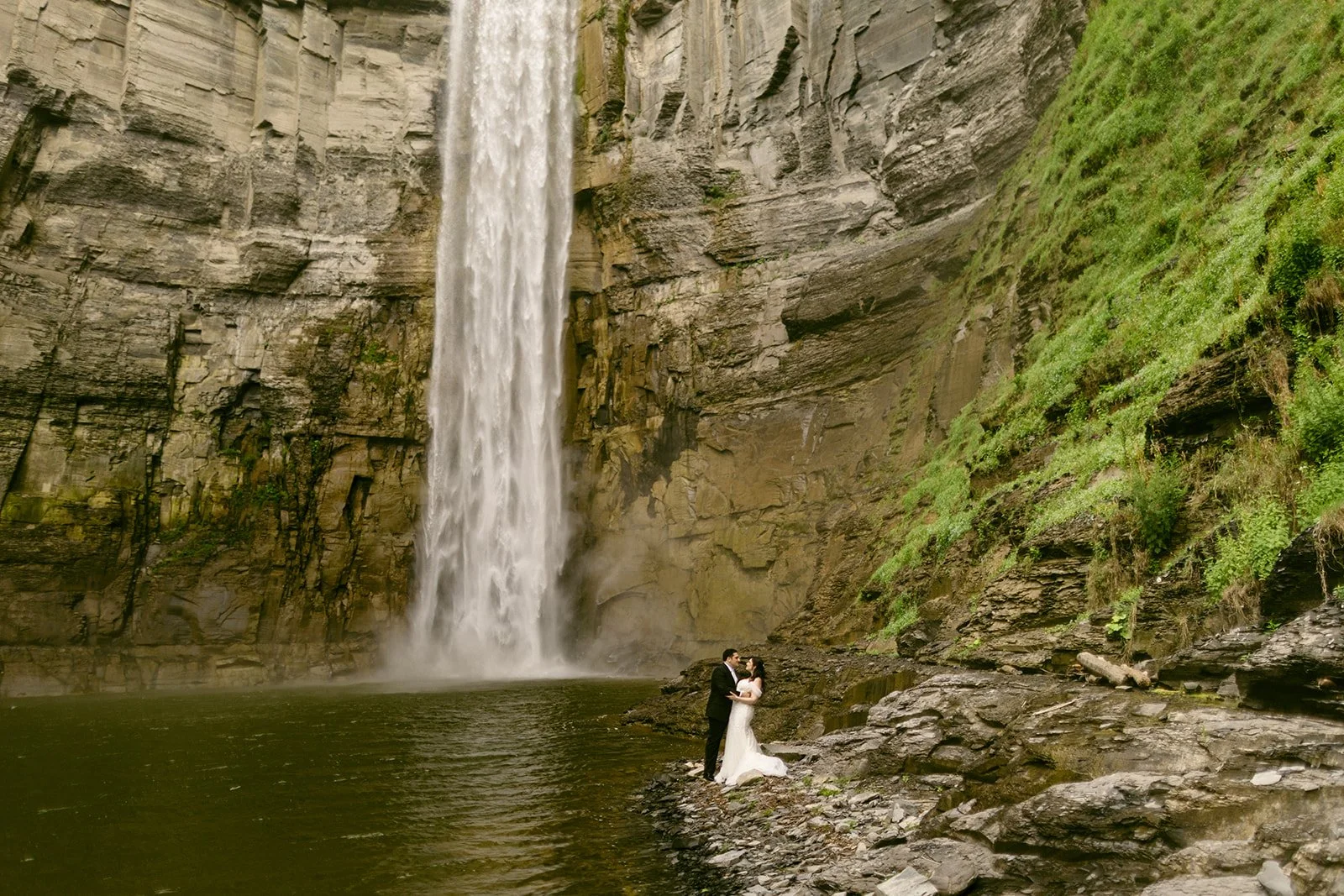 A bride and groom stand together near the base of a tall waterfall, surrounded by rocky cliffs and greenery at Taughannock Falls