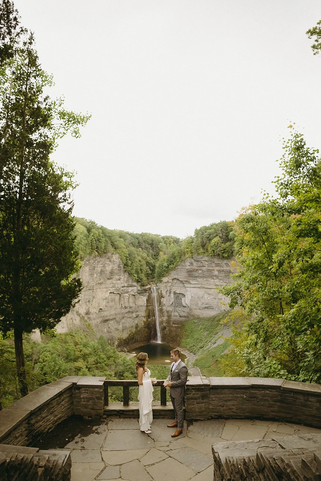 A bride and groom stand on a stone terrace overlooking a waterfall and cliffs for a wedding at Inn at Taughannock Falls Wedding