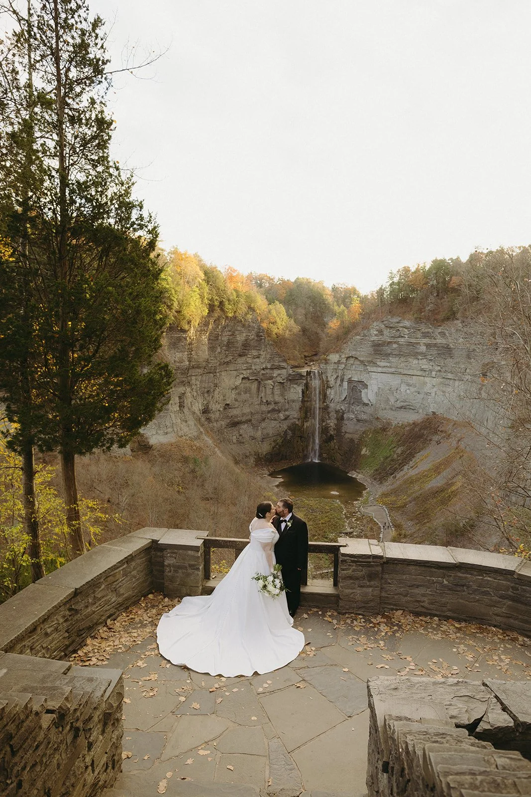 A bride and groom stand on a stone terrace overlooking a waterfall and cliffs for a wedding at Inn at Taughannock Falls Wedding