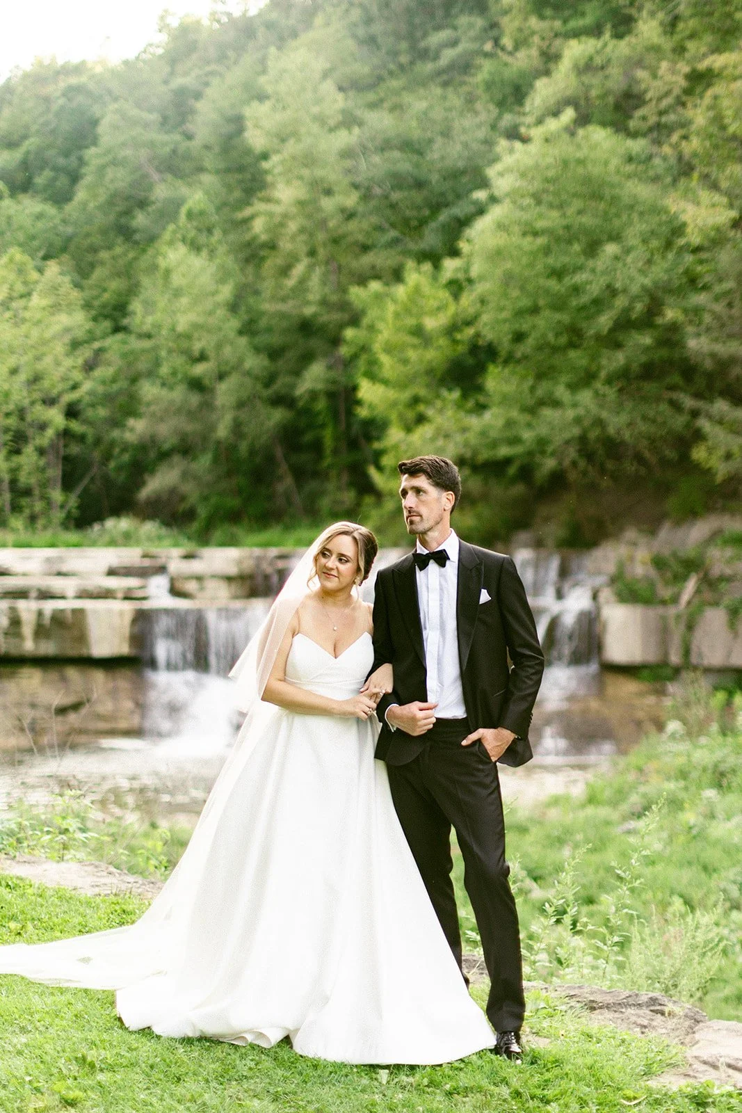 A bride in a white dress and a groom in a black suit stand together outdoors in front of a waterfall and greenery.