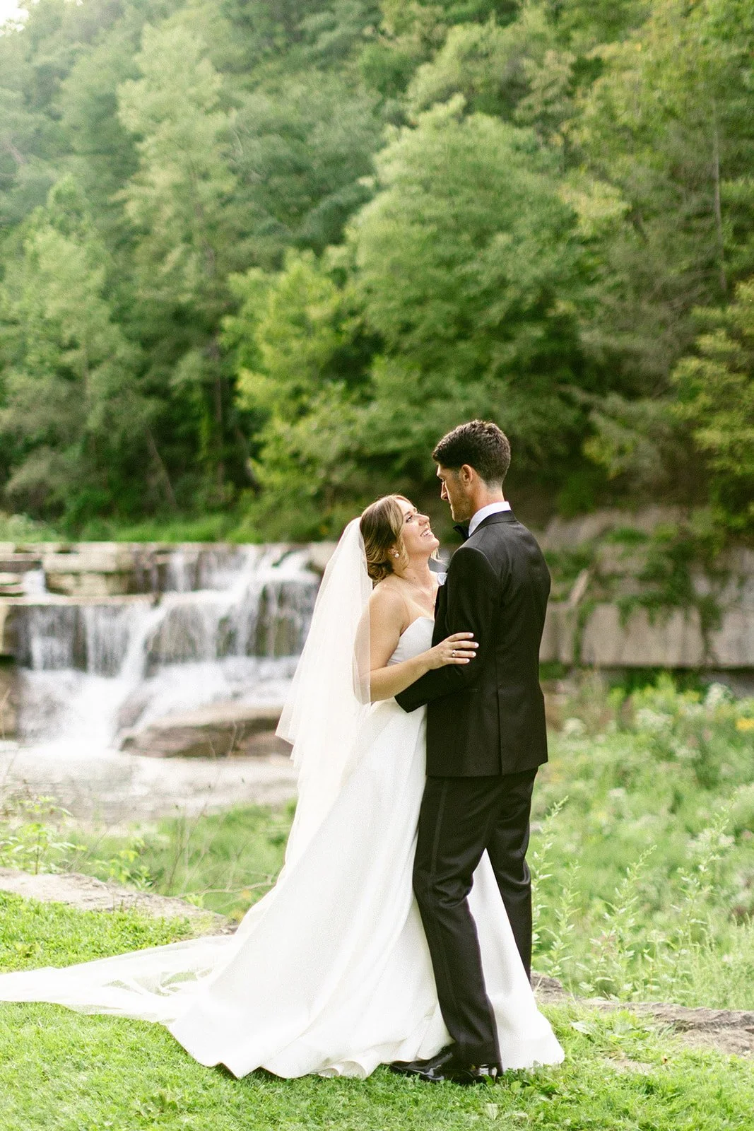 A bride in a white dress and a groom in a black suit stand together outdoors in front of a waterfall and greenery.