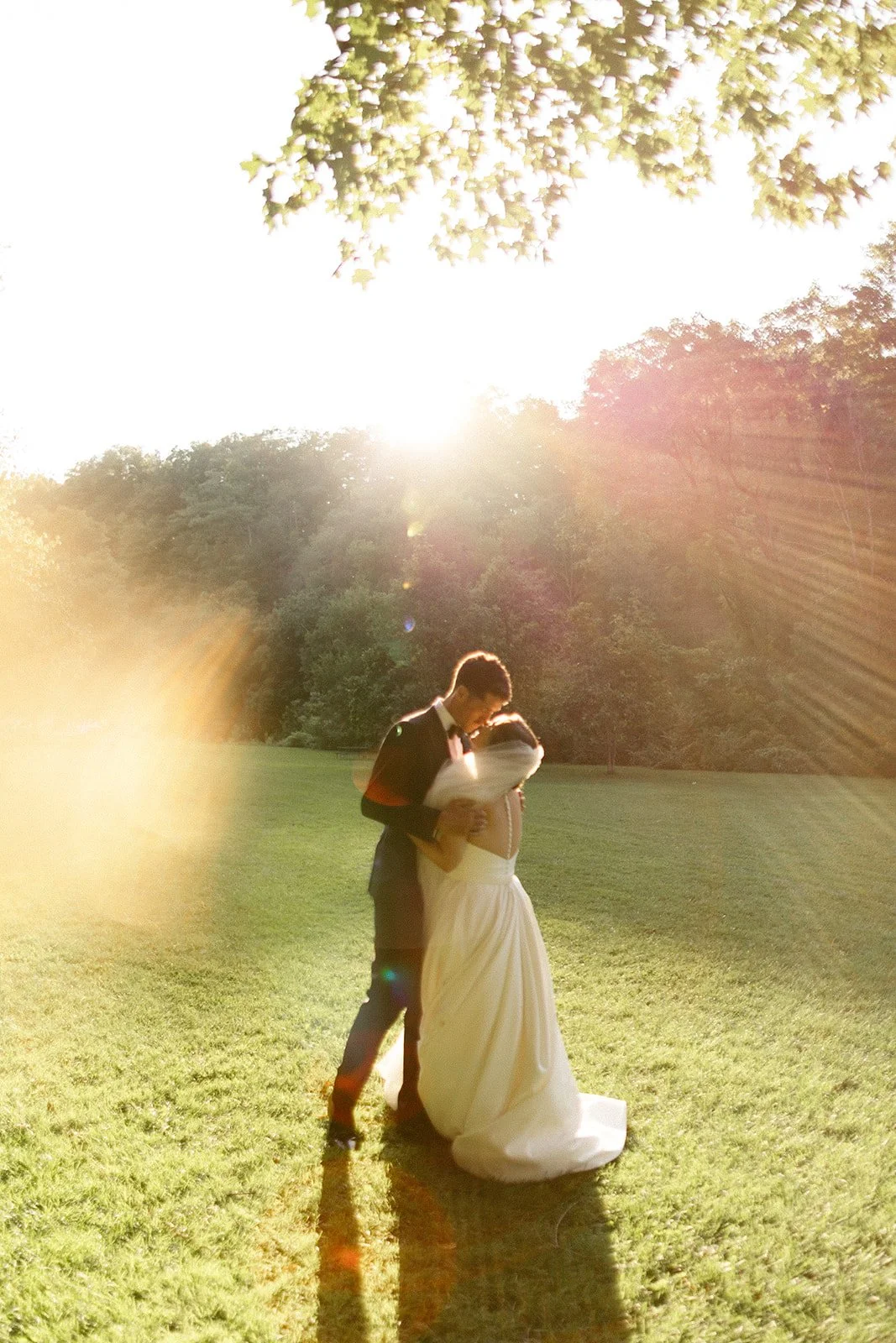 A bride in a white dress and a groom in a black suit stand together outdoors in front of a waterfall and greenery.