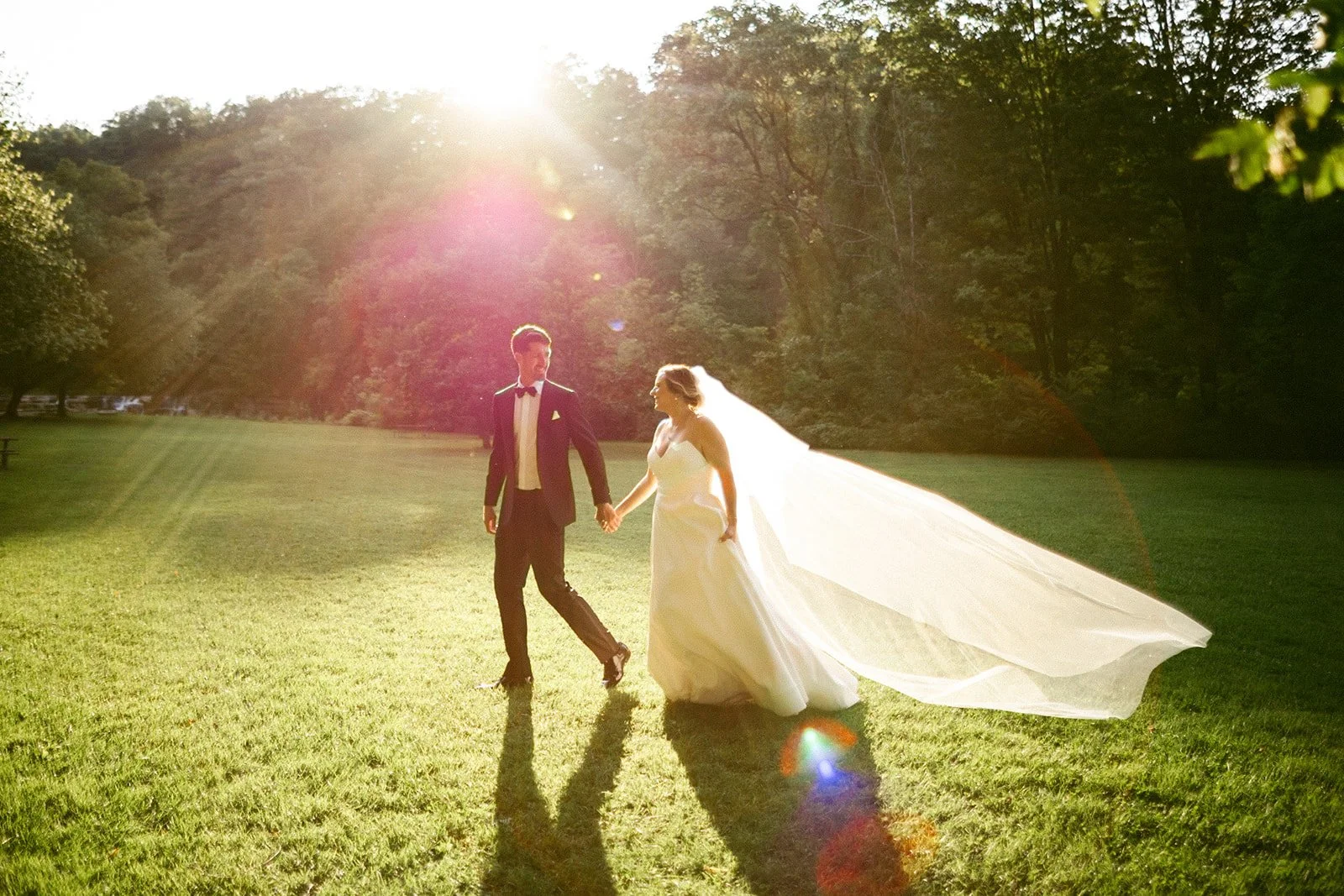 A bride and groom walk hand in hand on a grassy field at sunset, with trees in the background and sunlight shining through.