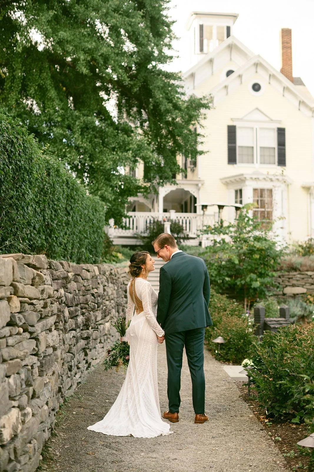 A bride and groom stand holding hands on a garden path, facing each other at the Inn at Taughannock Falls