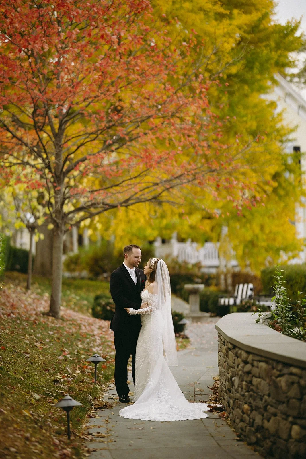 A bride and groom stand together on a walkway surrounded by autumn trees, with colorful leaves on the ground and a stone wall nearby