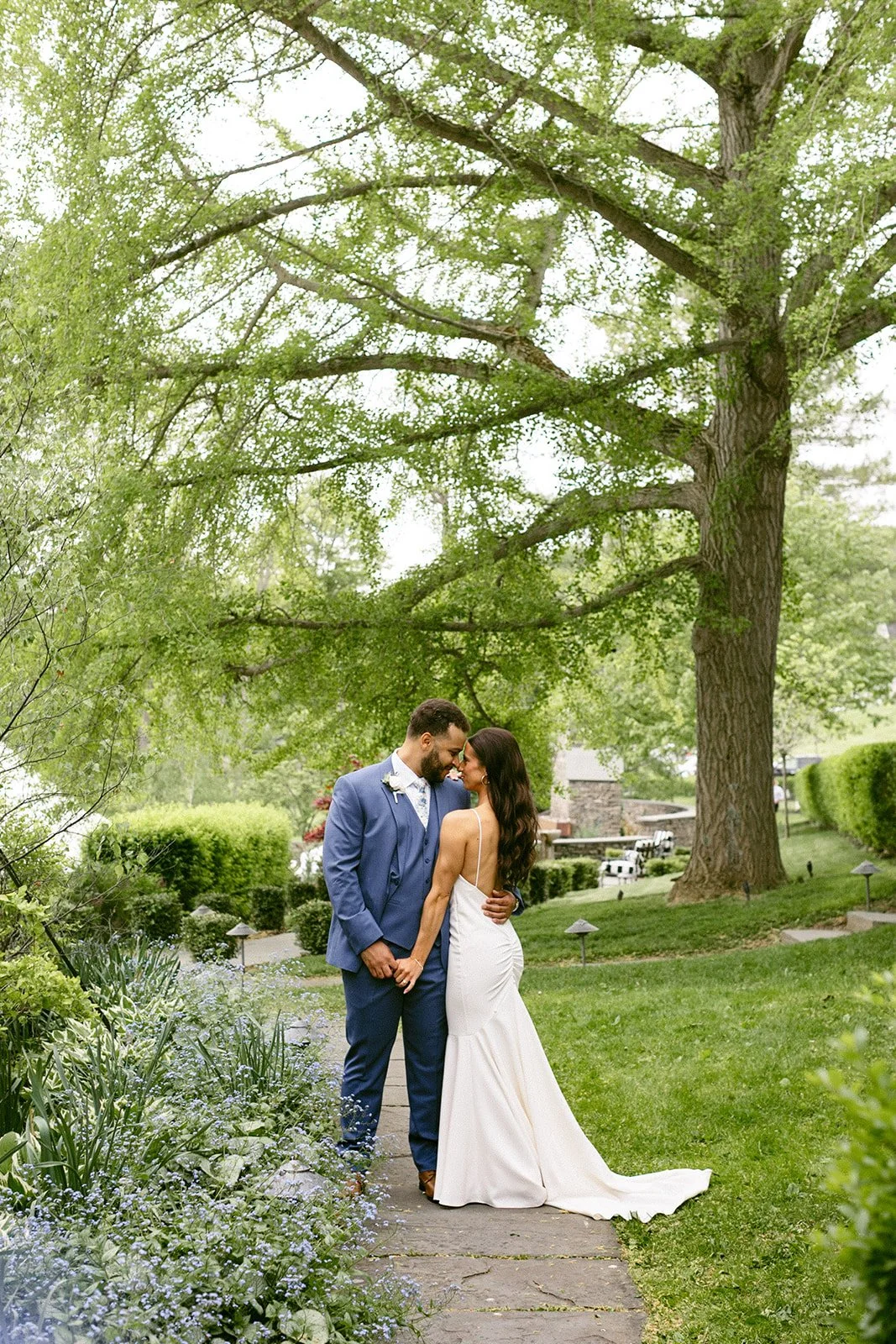 A bride and groom stand together on a garden path, embracing and smiling at each other with greenery and a large tree in the background.