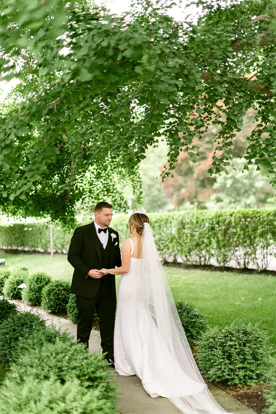 A bride and groom stand together on a large grassy lawn with trees and a white house in the background.