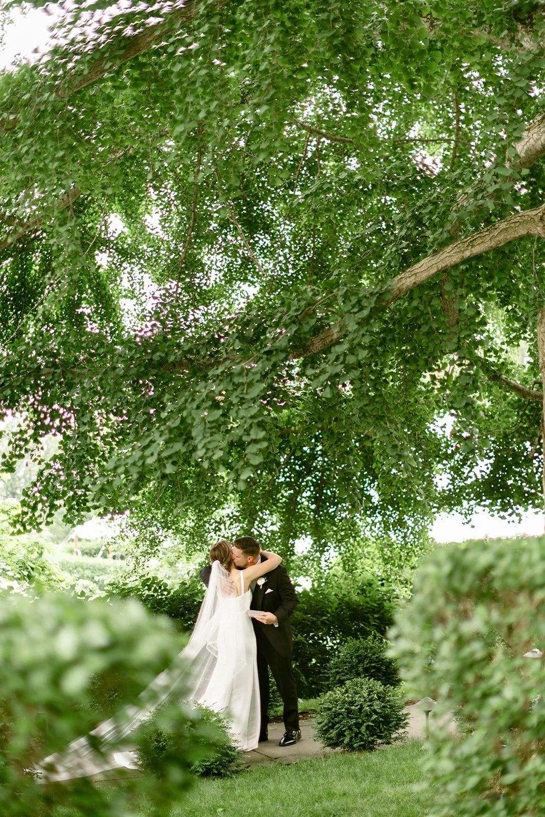 A bride and groom stand together on a large grassy lawn with trees and a white house in the background.