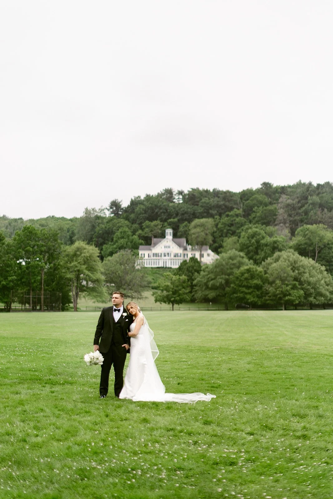 A bride and groom stand together on a large grassy lawn with trees and a white house in the background.