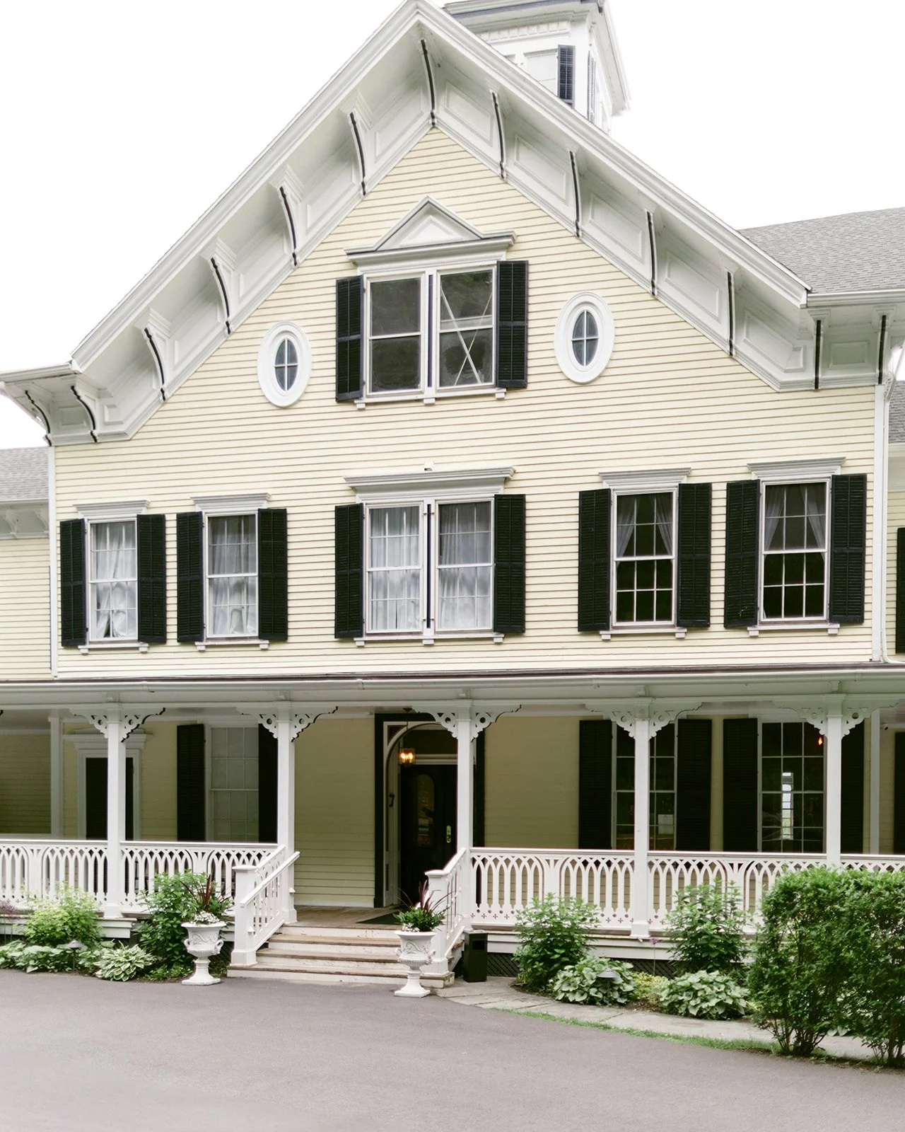 Large white house with multiple gables, a sunroom, and a small tower, surrounded by greenery and a neatly manicured lawn under a clear sky at the Inn at Taughannock Falls 