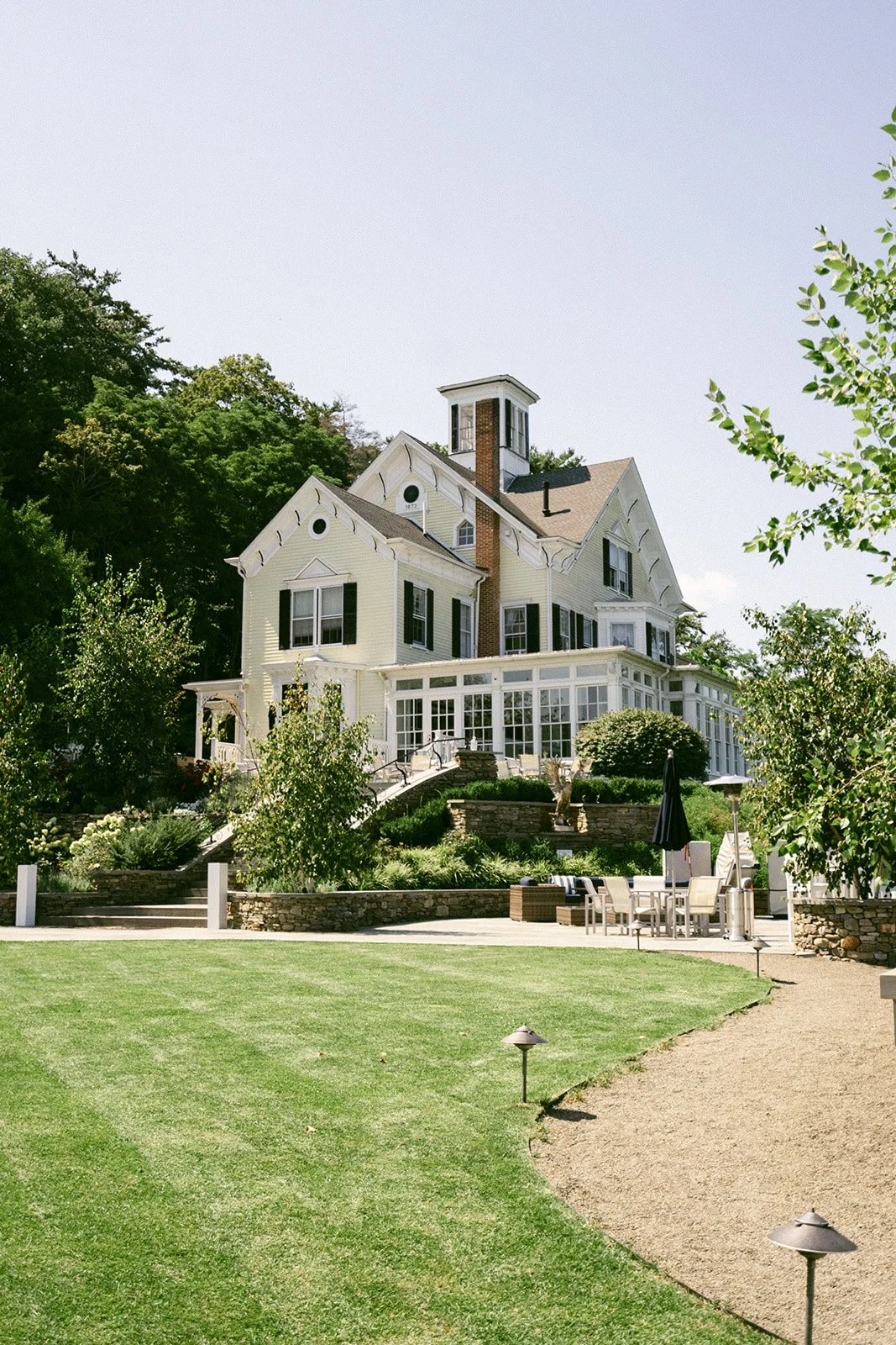 Large white house with multiple gables, a sunroom, and a small tower, surrounded by greenery and a neatly manicured lawn under a clear sky at the Inn at Taughannock Falls 