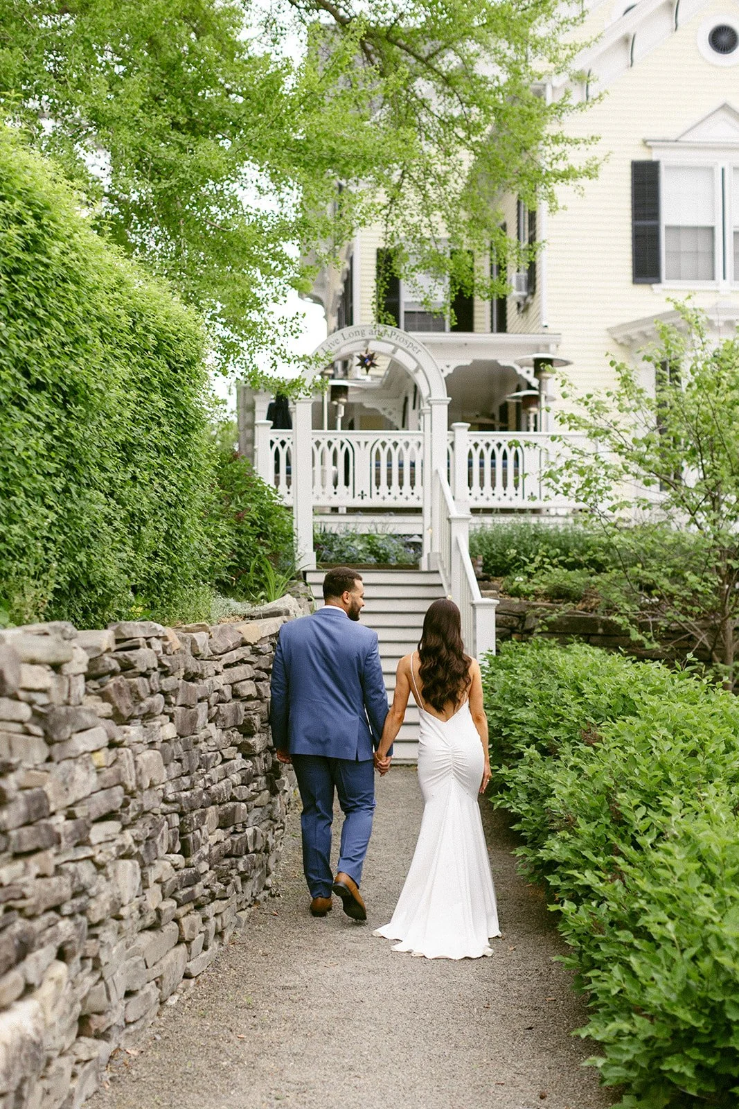 A newlywed couple stands side by side on a stone path in front of a white house, posing for a wedding photo. The groom wears a blue suit; the bride wears a white dress and holds a bouquet.