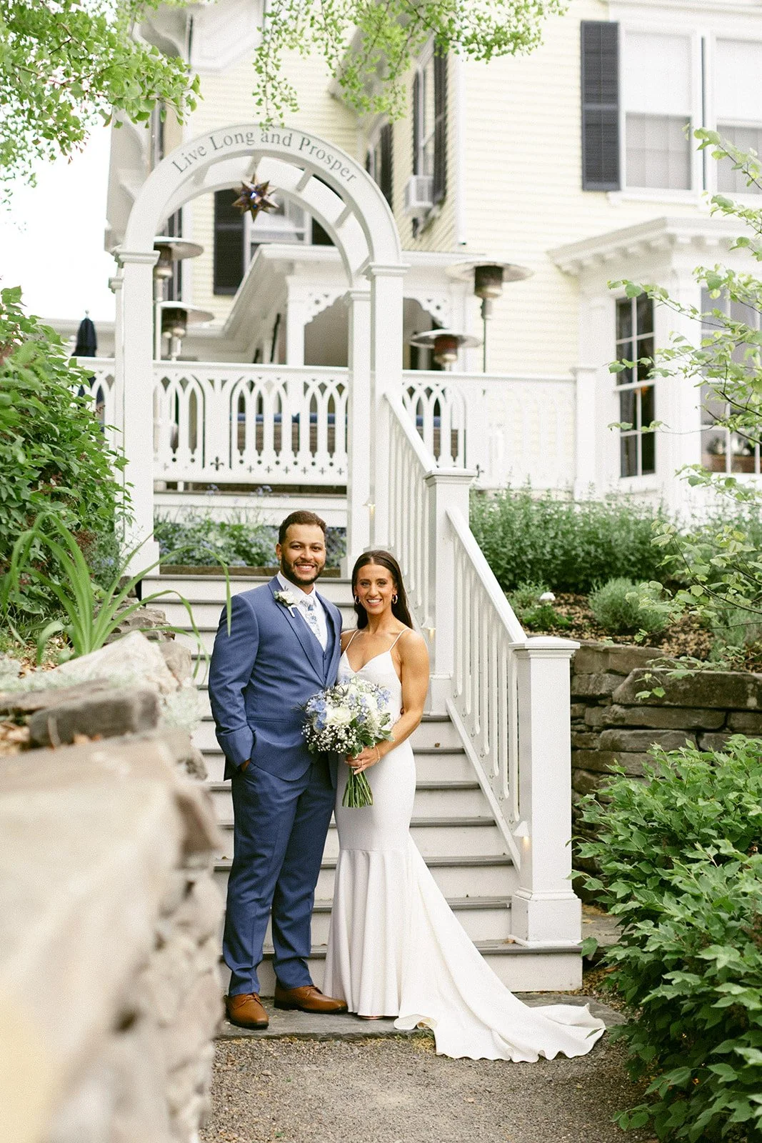 A newlywed couple stands side by side on a stone path in front of a white house, posing for a wedding photo. The groom wears a blue suit; the bride wears a white dress and holds a bouquet.