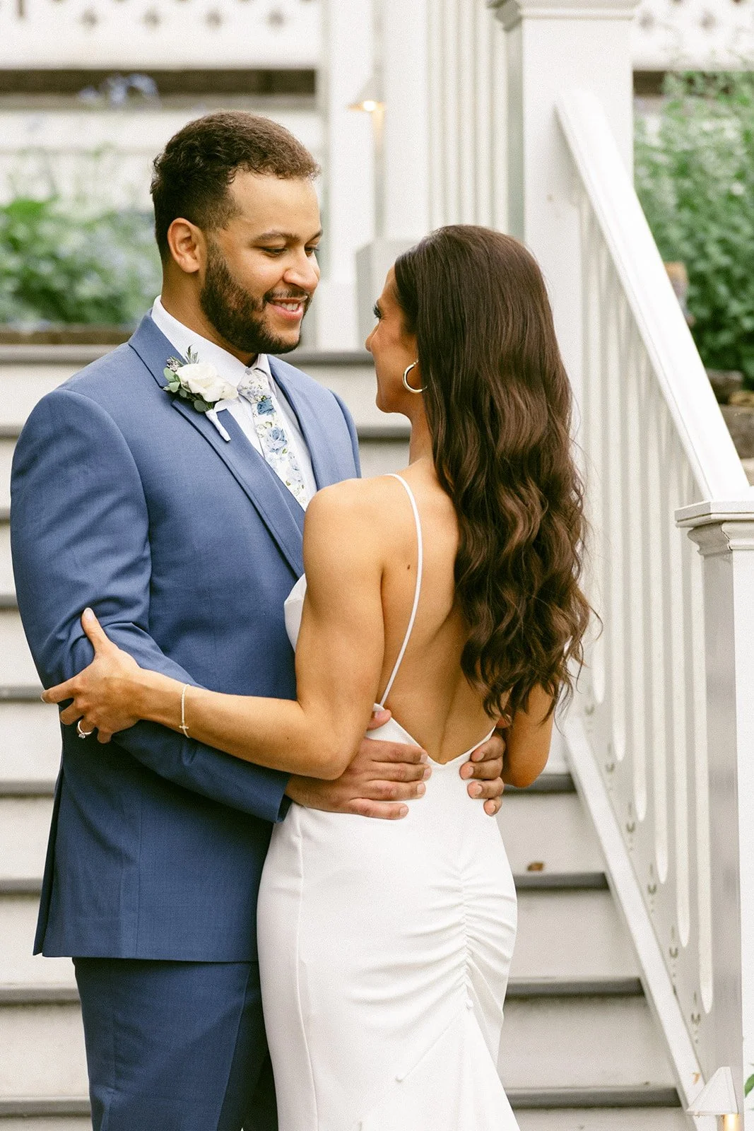 A newlywed couple stands side by side on a stone path in front of a white house, posing for a wedding photo. The groom wears a blue suit; the bride wears a white dress and holds a bouquet.