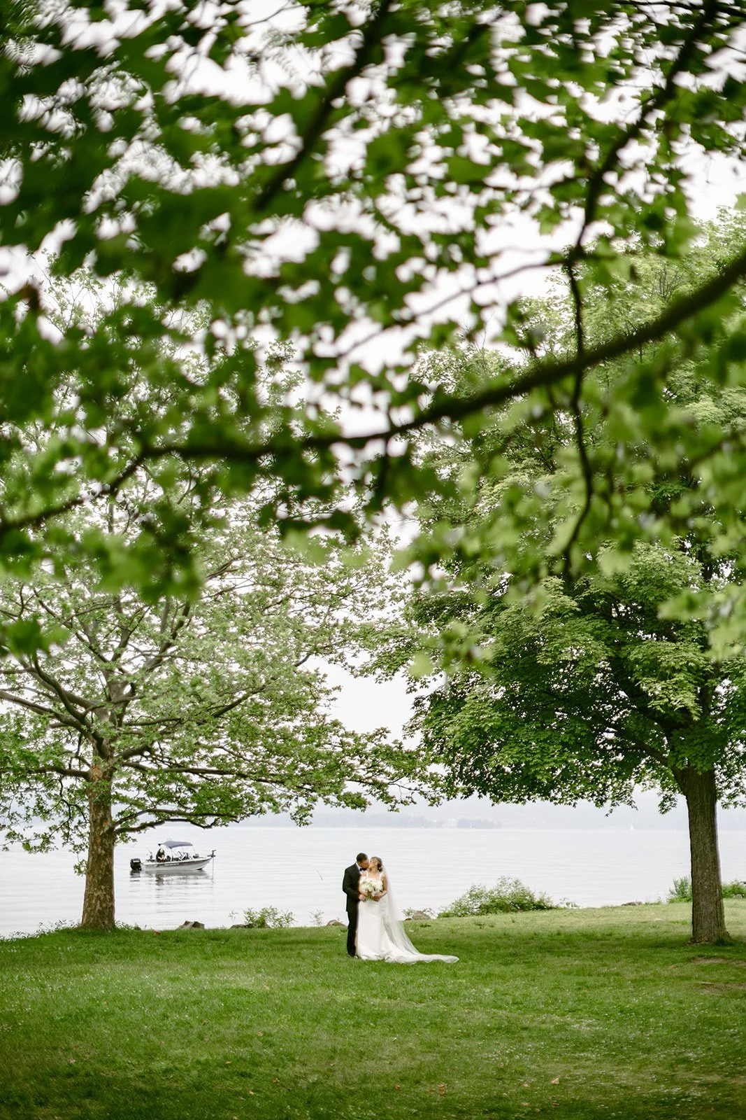 A bride and groom stand together on grass between two large trees, with a body of water visible in the background.