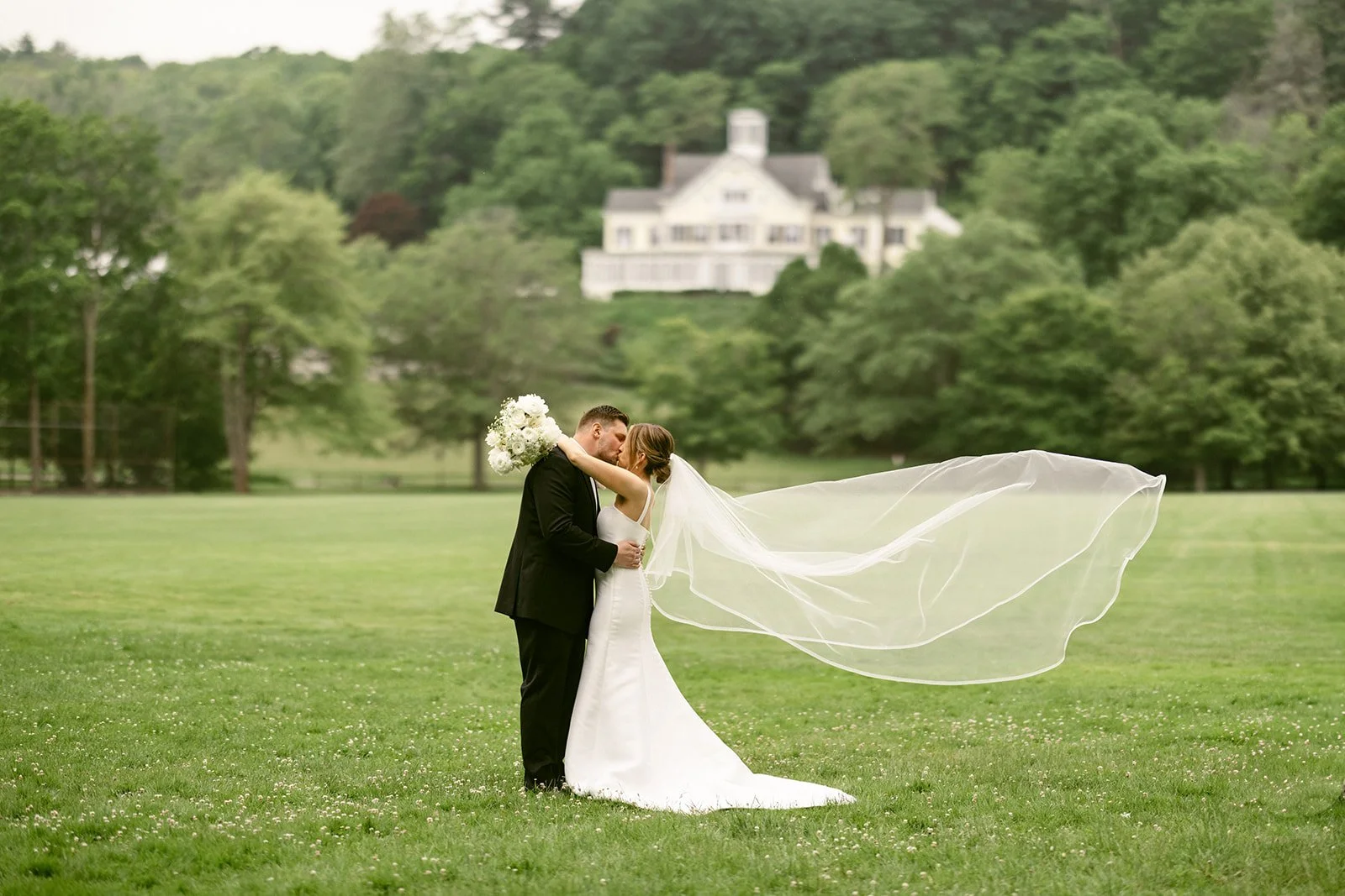 A bride and groom stand together on grass between two large trees, with a body of water visible in the background.