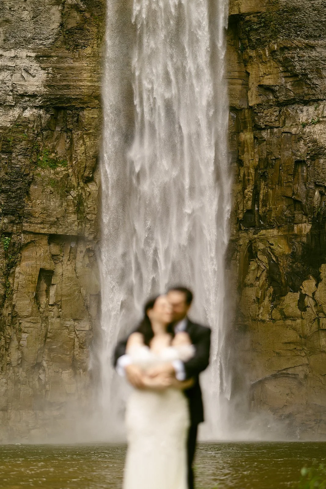 A bride and groom stand together near the base of a tall waterfall, surrounded by rocky cliffs and greenery at Taughannock Falls
