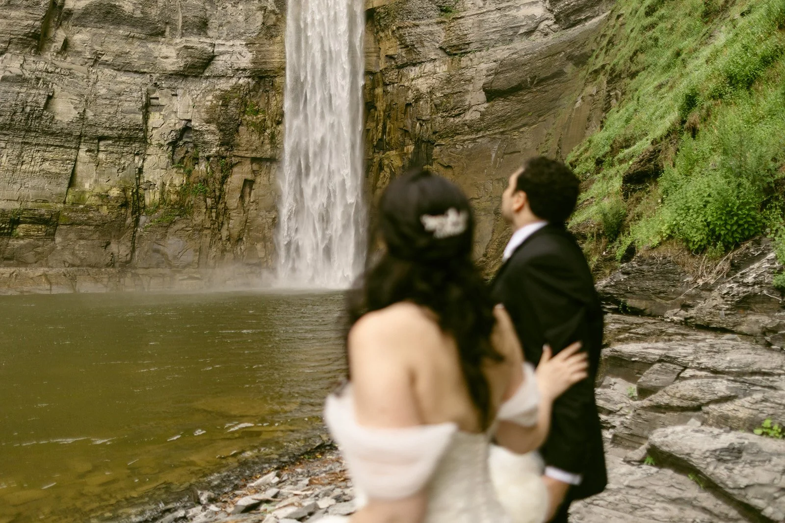 A bride and groom stand together near the base of a tall waterfall, surrounded by rocky cliffs and greenery at Taughannock Falls