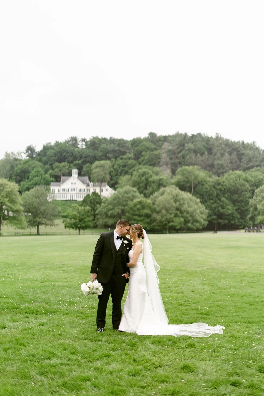 A bride and groom stand together on grass between two large trees, with a body of water visible in the background.
