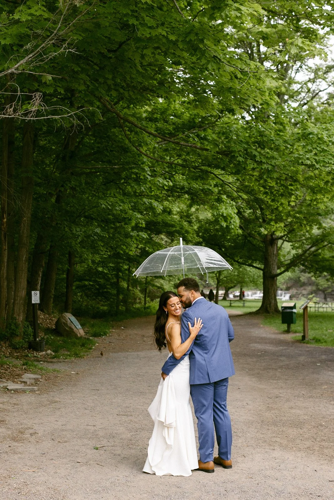 A couple dressed in wedding attire stands on a tree-lined path, embracing under a clear umbrella on a cloudy day.
