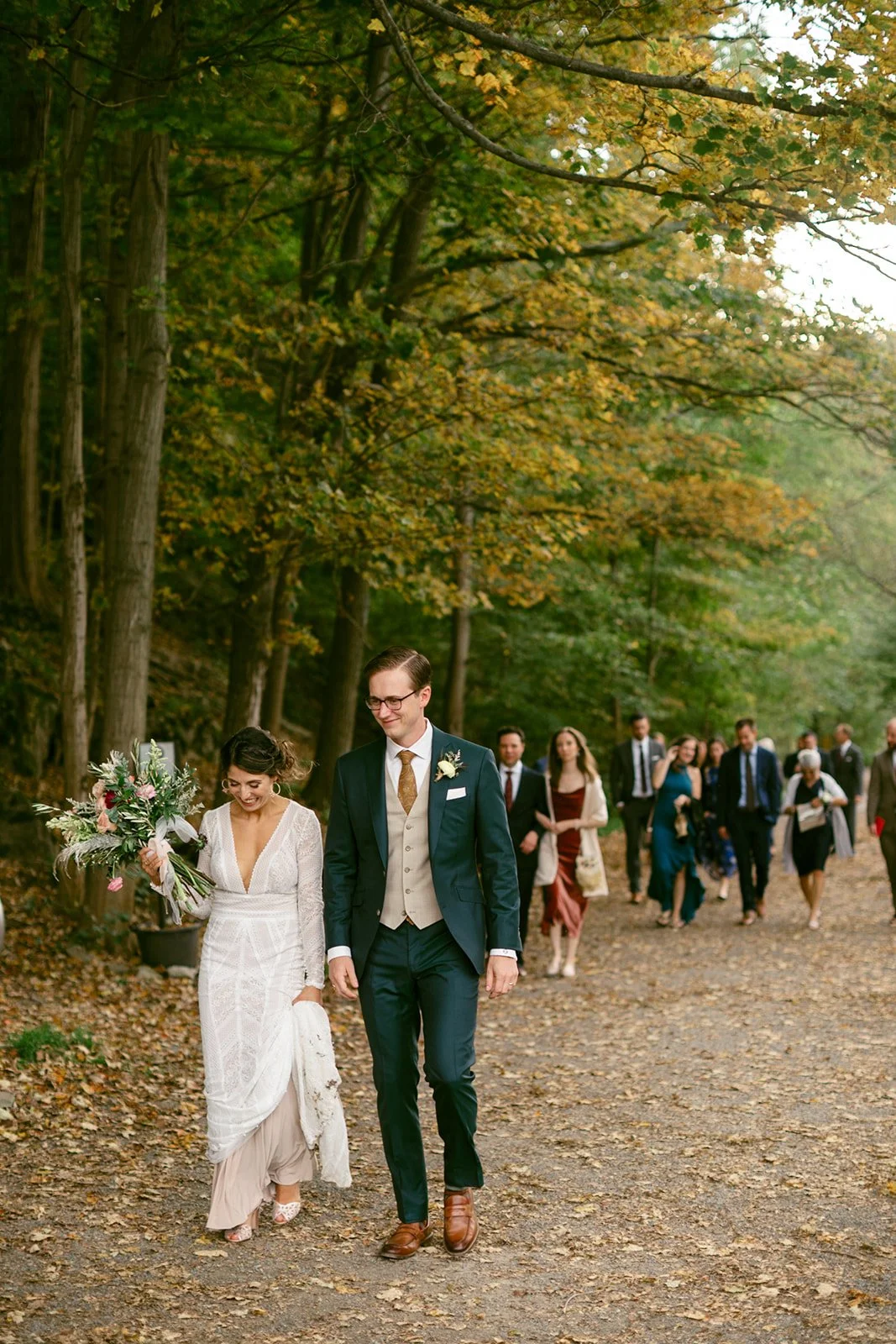 A bride and groom walk along a wooded path, followed by a group of well-dressed guests on a forested trail surrounded by autumn leaves.