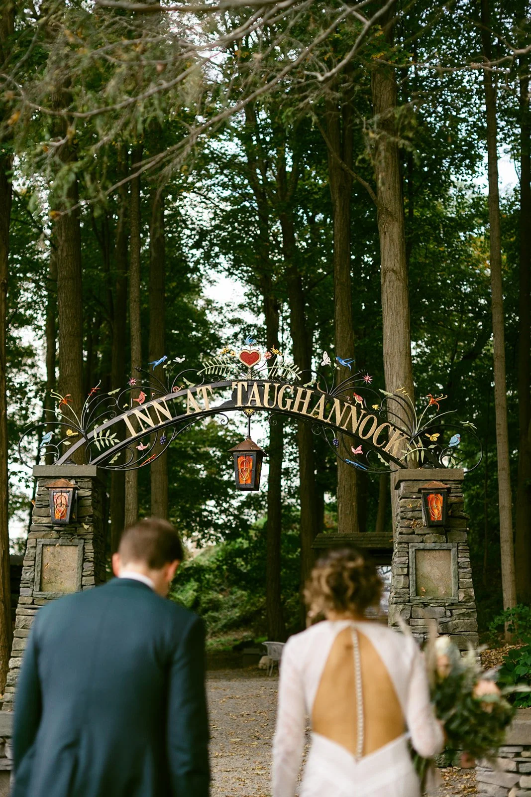 A couple in formal attire walks toward an arched entrance sign reading "The Inn at Taughannock" in a wooded outdoor setting.