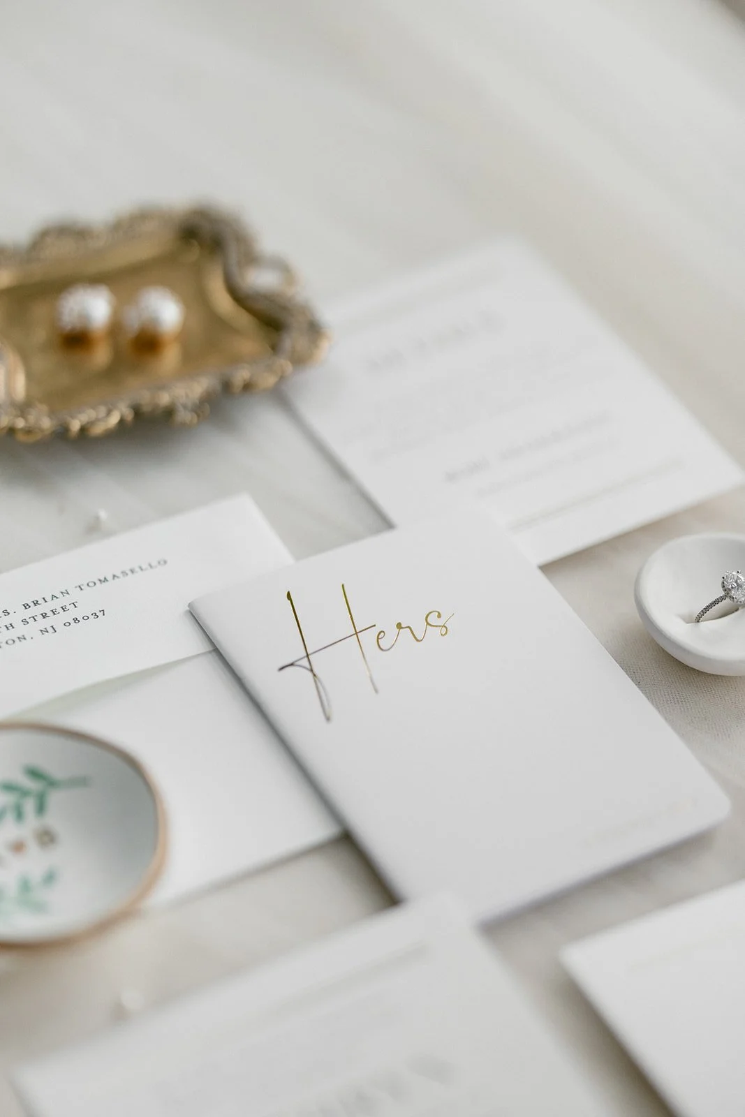 A flat lay of wedding stationery, including a booklet labeled "Hers," a dish with rings, a tray holding pearl earrings, and other white cards arranged on a light surface.