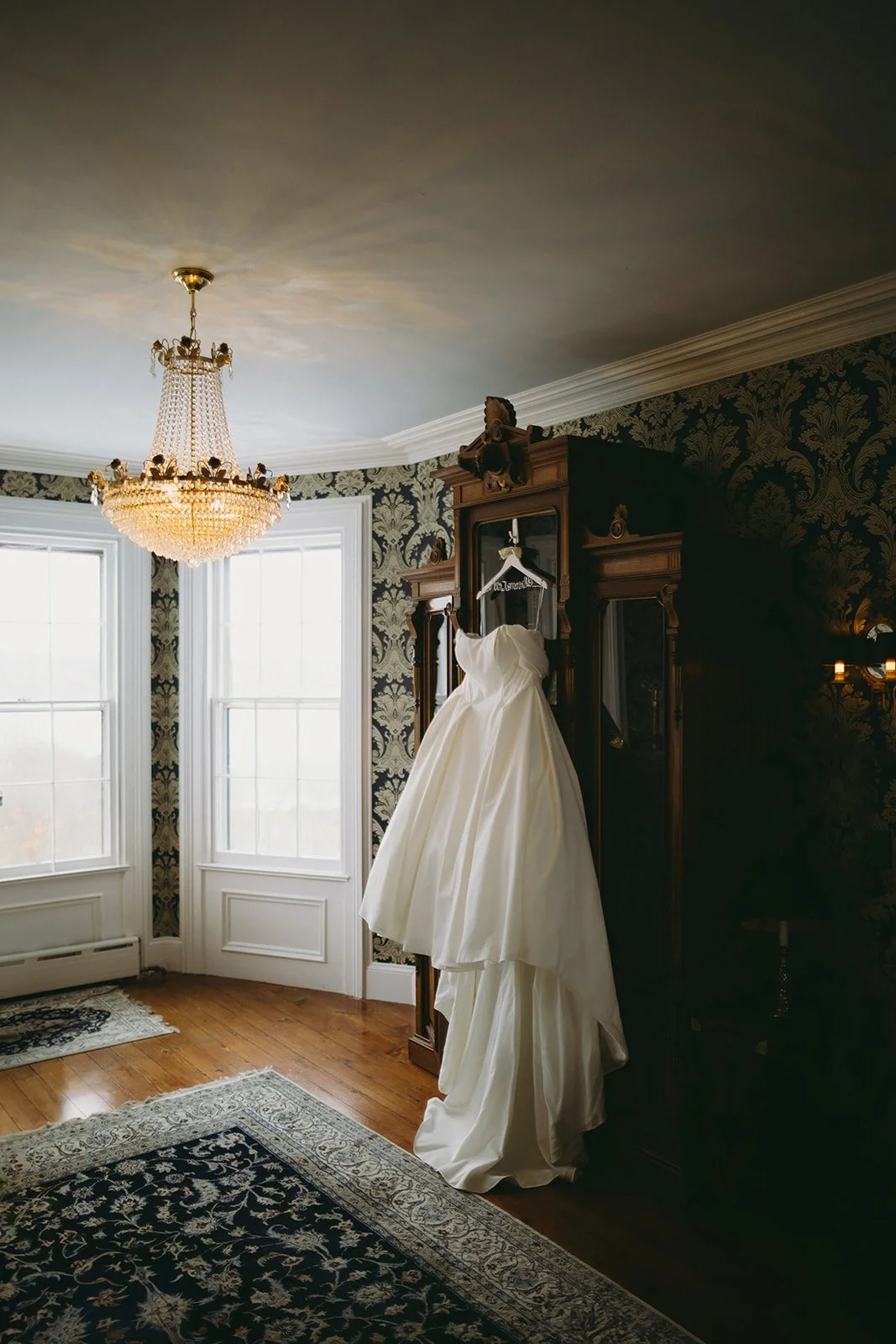 A white wedding dress hangs on a wooden wardrobe in a room with patterned wallpaper, large windows, a chandelier, and decorative rugs.