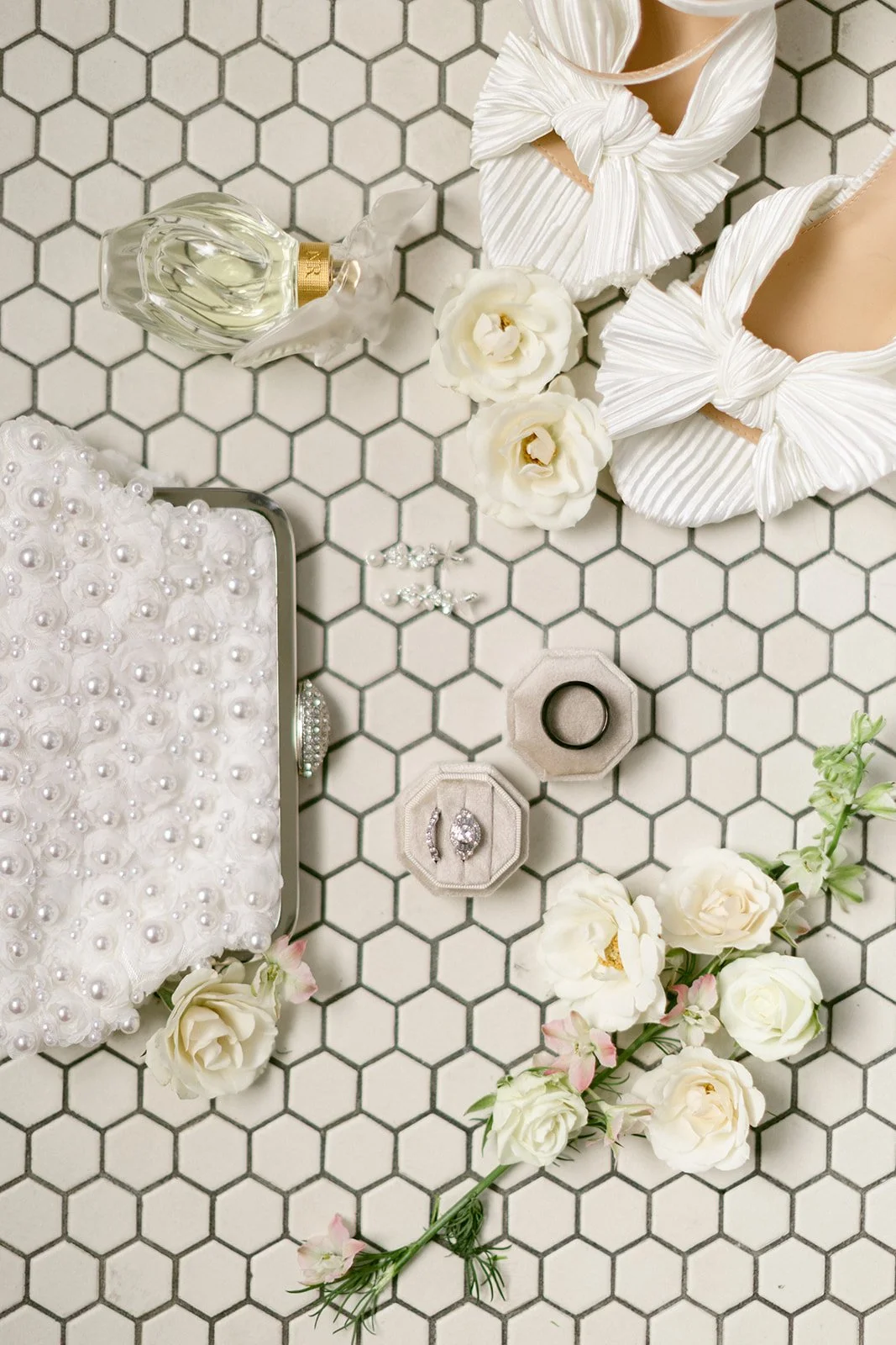 Flat lay of bridal accessories on hexagonal tile floor, including white shoes, pearl clutch, ring box, perfume bottle, earrings, and white flowers.