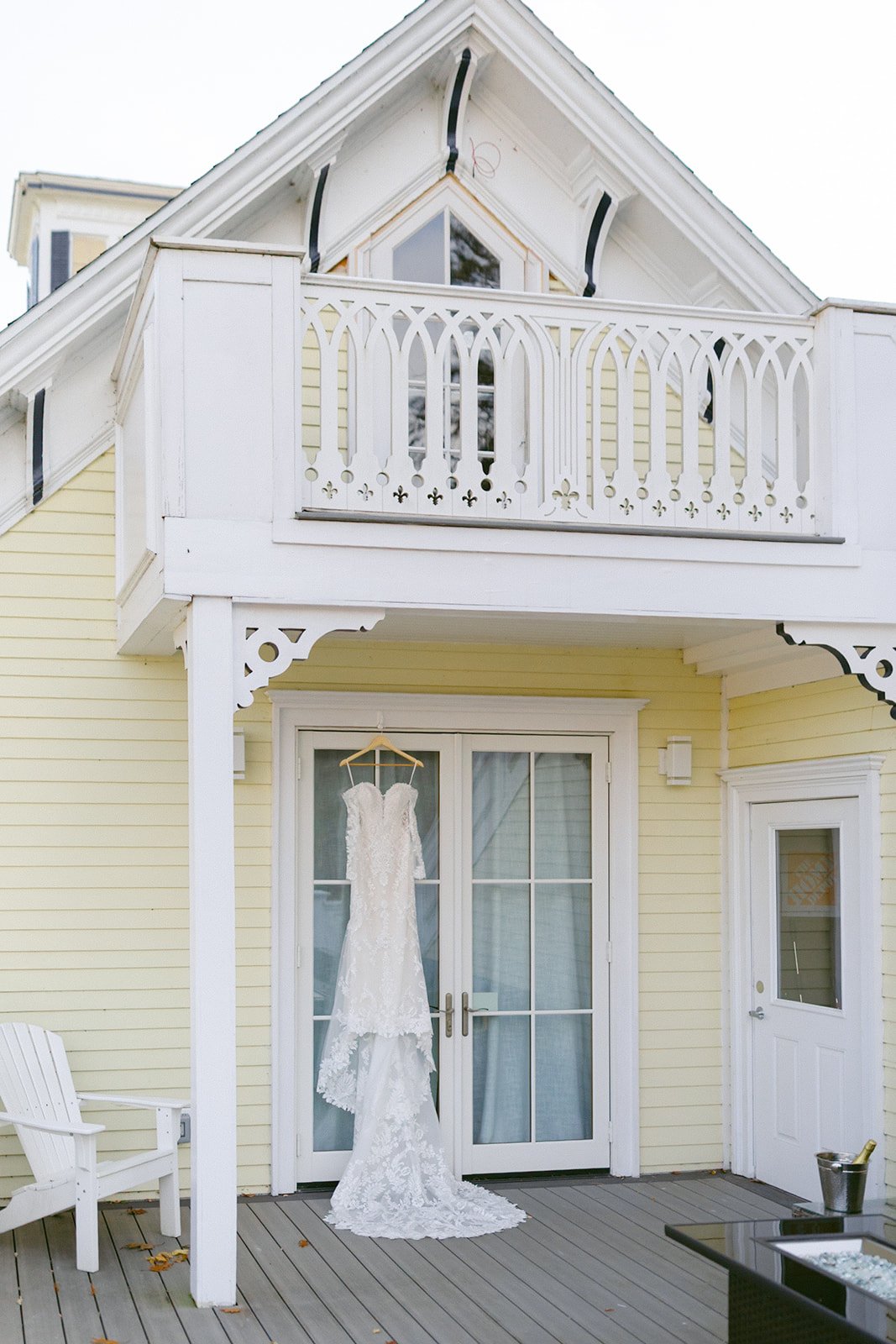 A white lace wedding dress hangs on the double doors of a light yellow house with a white balcony and decorative trim.