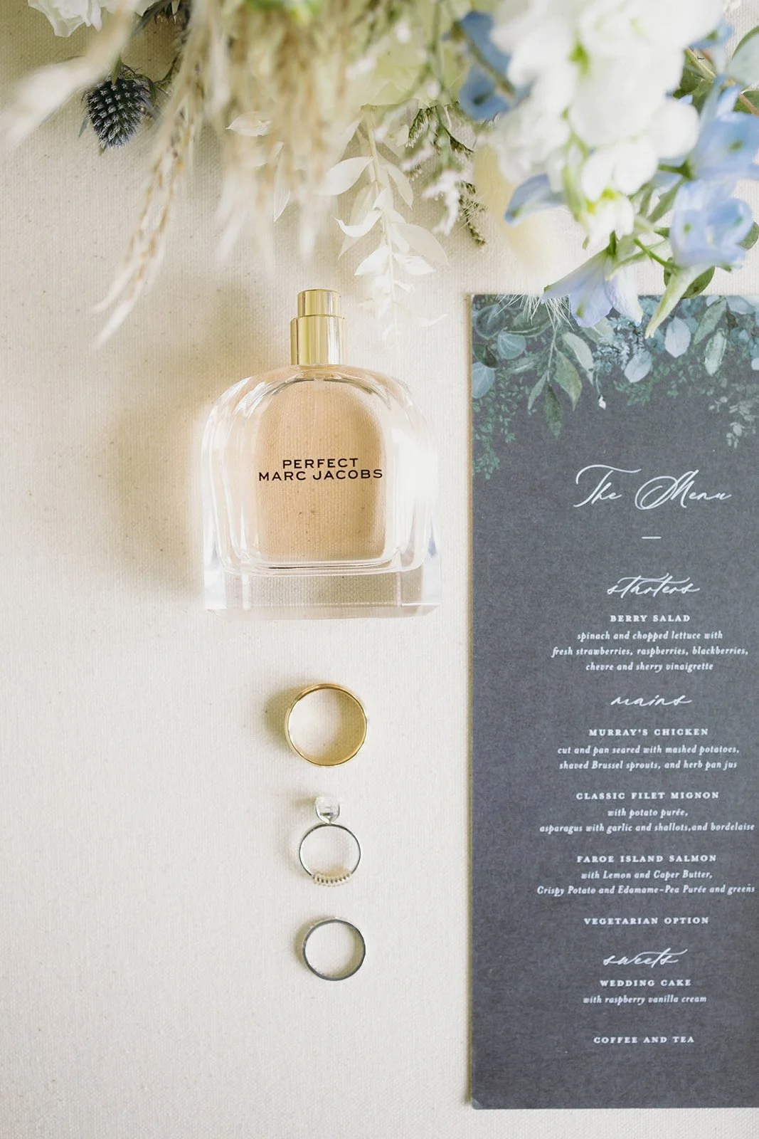 A bottle of Perfect Marc Jacobs perfume, three rings, and a printed menu are arranged on a light surface with flowers at the top.