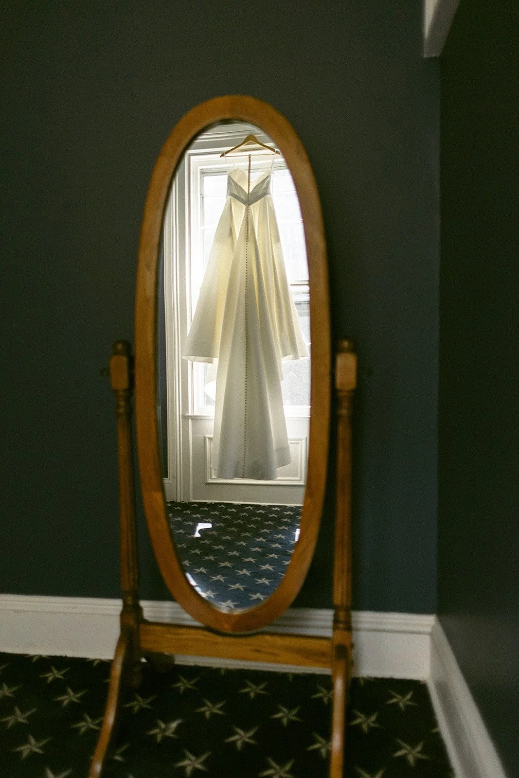 A white lace wedding dress hangs on a wooden hanger in front of a window framed by dark curtains in a dimly lit room.
