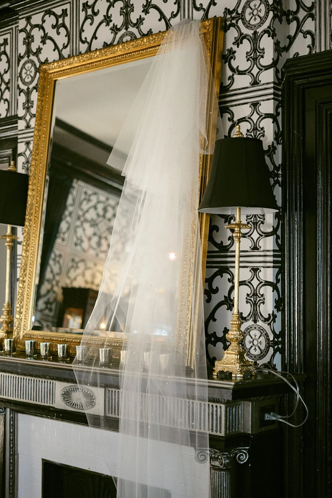 A gold-framed mirror above a mantel with black-and-white patterned wallpaper, two black-shaded lamps, and a white veil draped over the mirror.