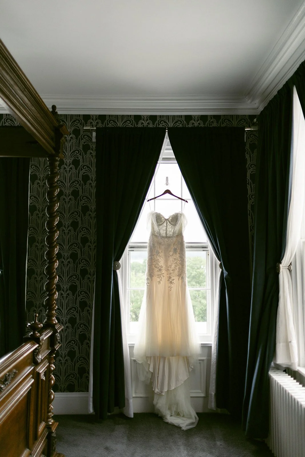 A white lace wedding dress hangs on a wooden hanger in front of a window framed by dark curtains in a dimly lit room.