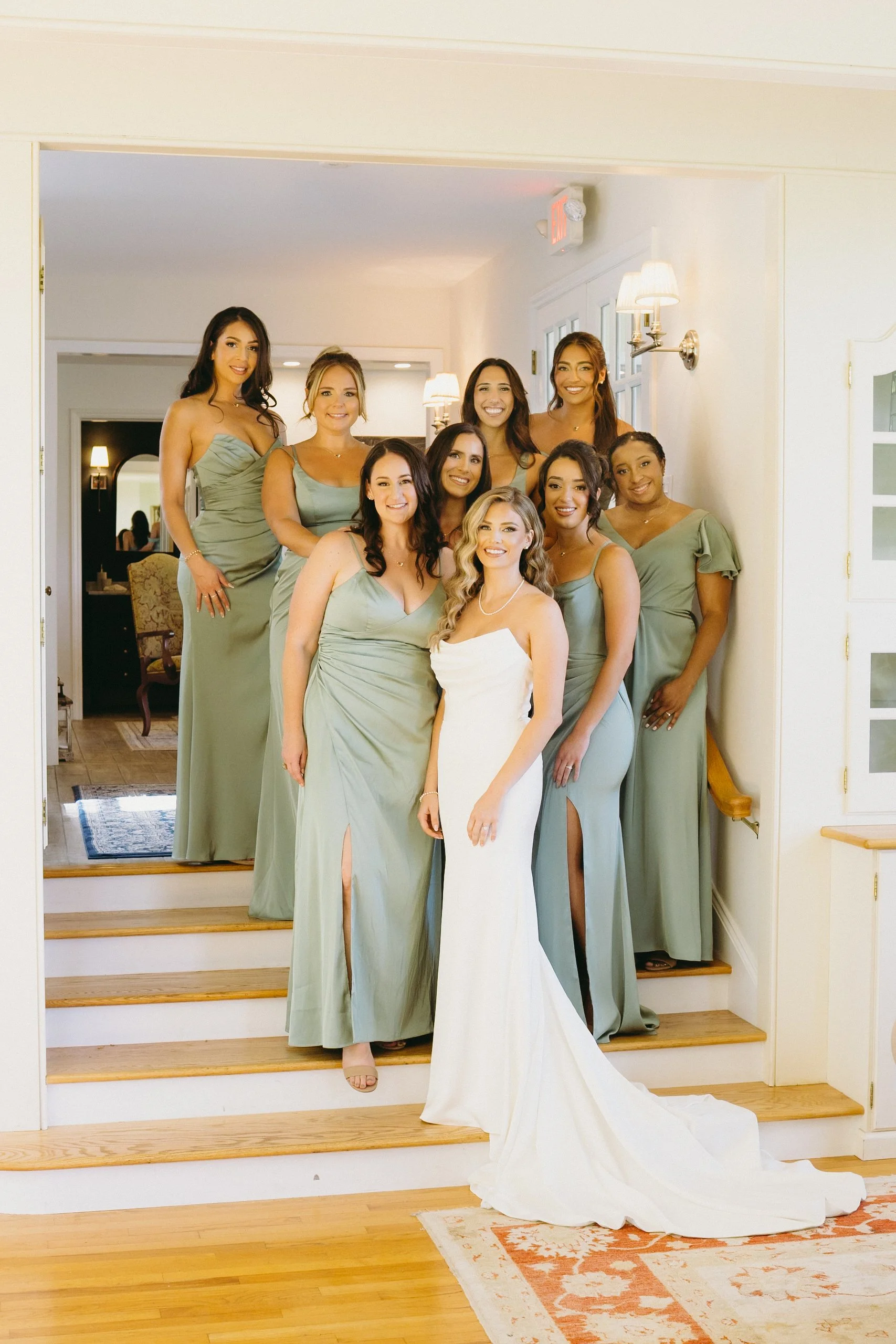 A bride in a white dress stands with eight bridesmaids in sage green dresses on indoor steps, all posing for a group photo.