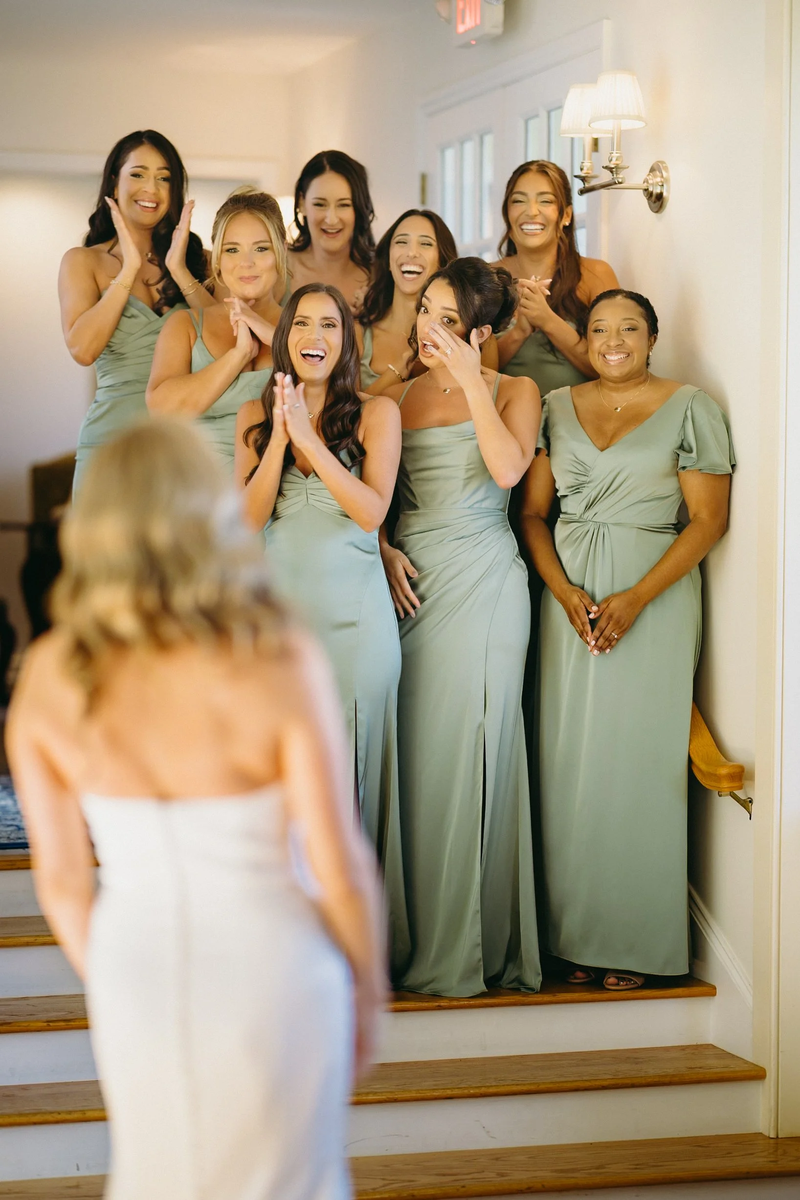 A bride in a white dress stands with eight bridesmaids in sage green dresses on indoor steps, all posing for a group photo.
