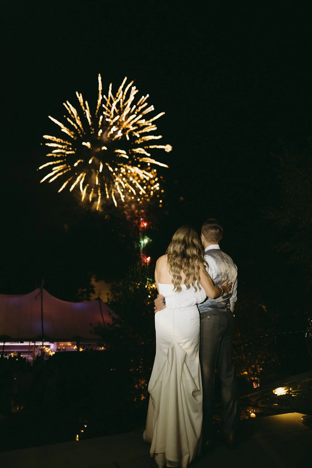 A couple in formal attire stands together at night, watching fireworks light up the dark sky.