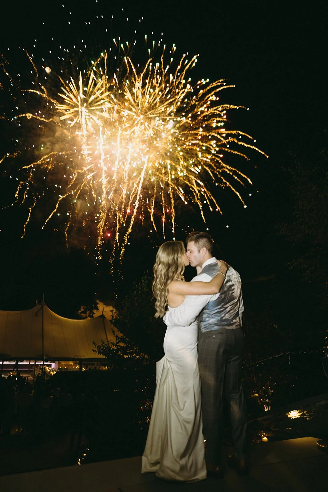 A couple in formal attire stands together at night, watching fireworks light up the dark sky.