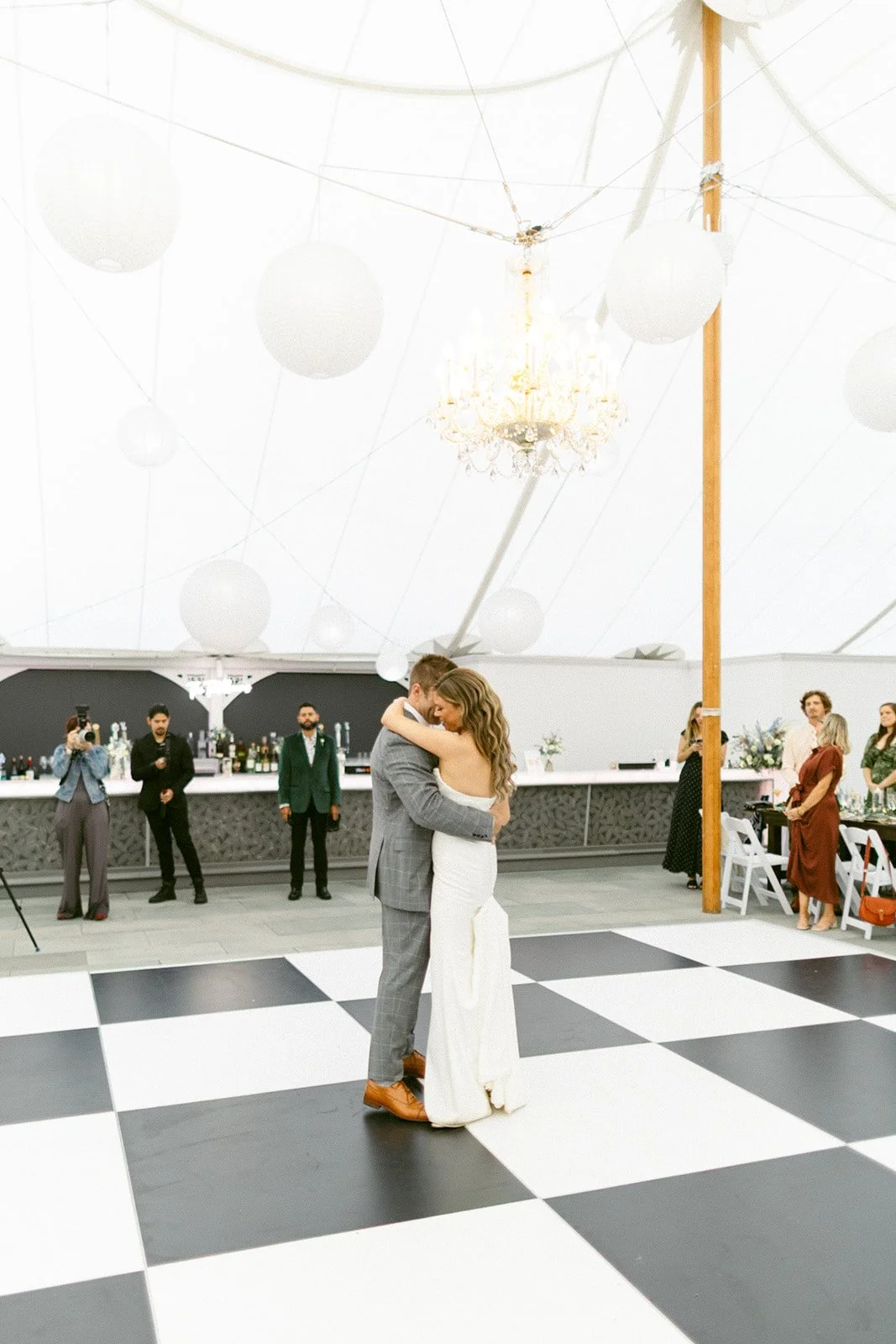 A bride and groom share their first dance on a black-and-white checkered floor under a white tent with a chandelier, while guests watch and take photos.