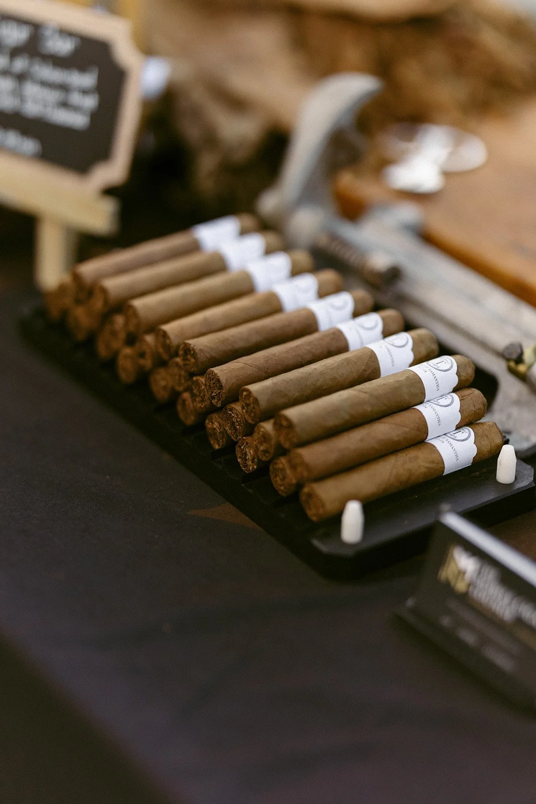 A row of cigars with white bands is neatly arranged on a black tray, displayed on a dark tablecloth.