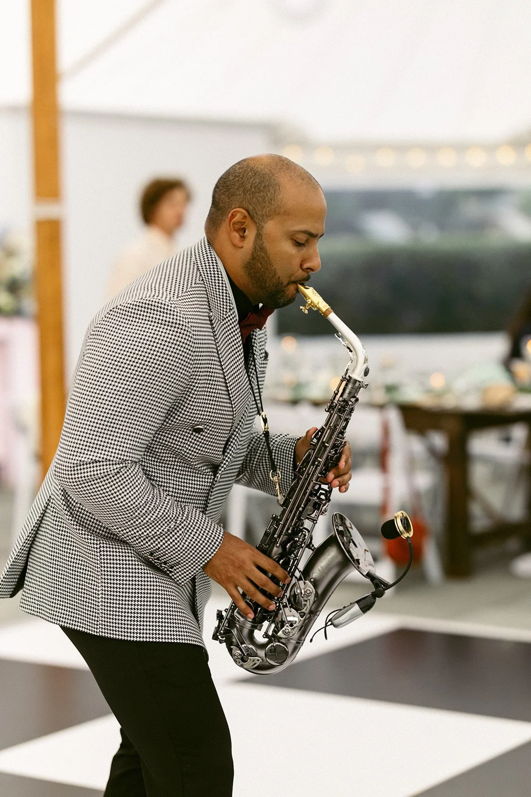 A man in a checkered jacket plays a saxophone indoors on a black and white checkered floor, with blurred people and tables in the background.