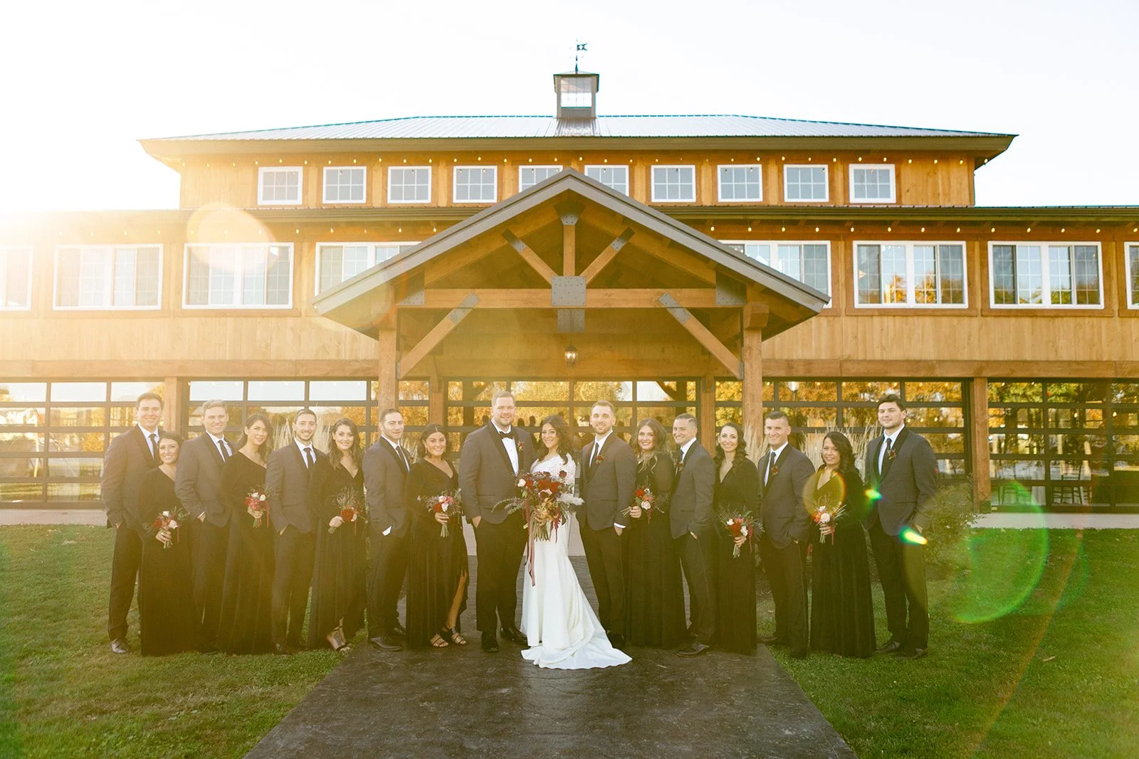  A wedding party poses in front of a large wooden barn-style building at Colloca Estate Winery an upstate ny wedding venue