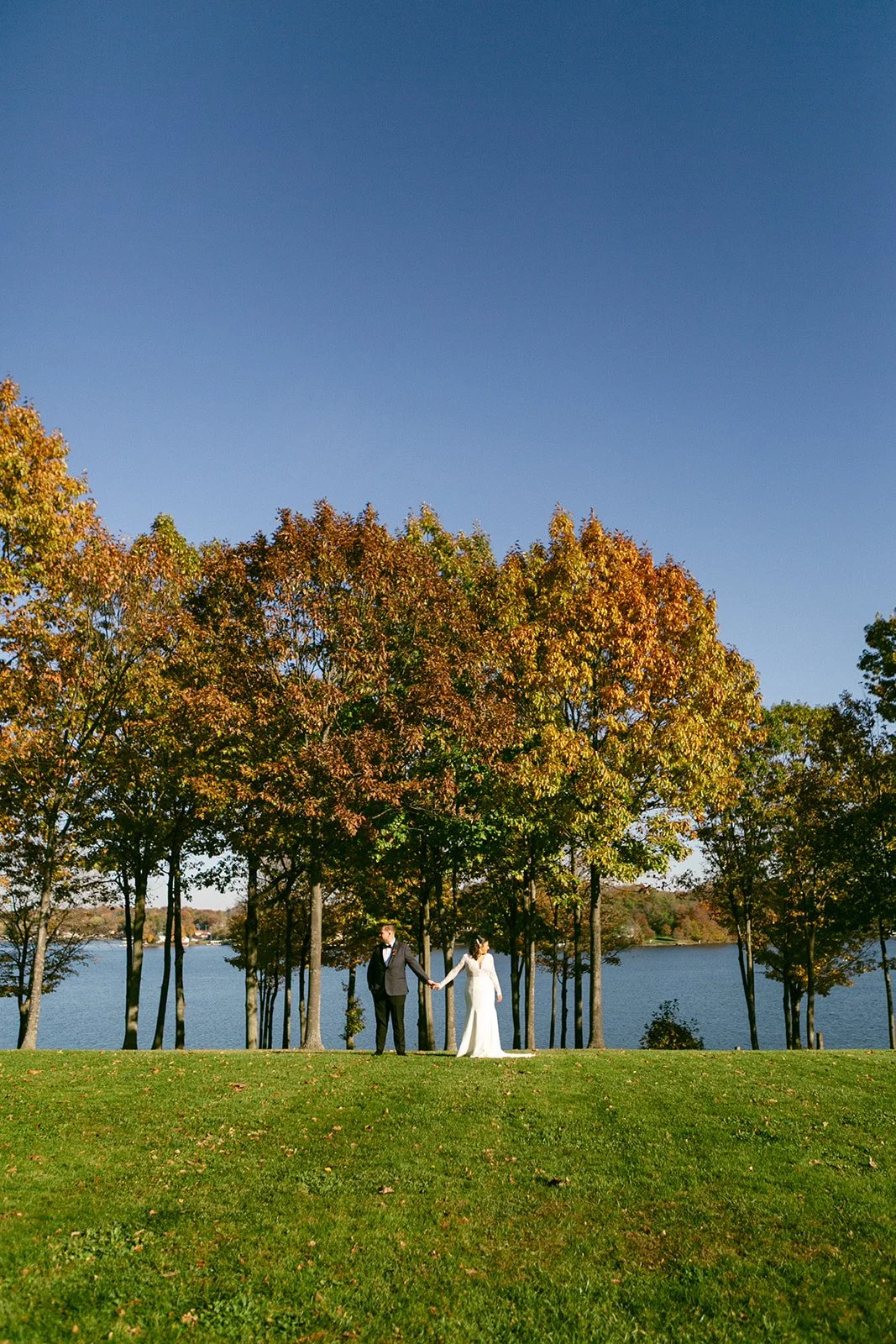A bride and groom hold hands on a grassy hill with autumn trees and a lake in the background under a clear blue sky.
