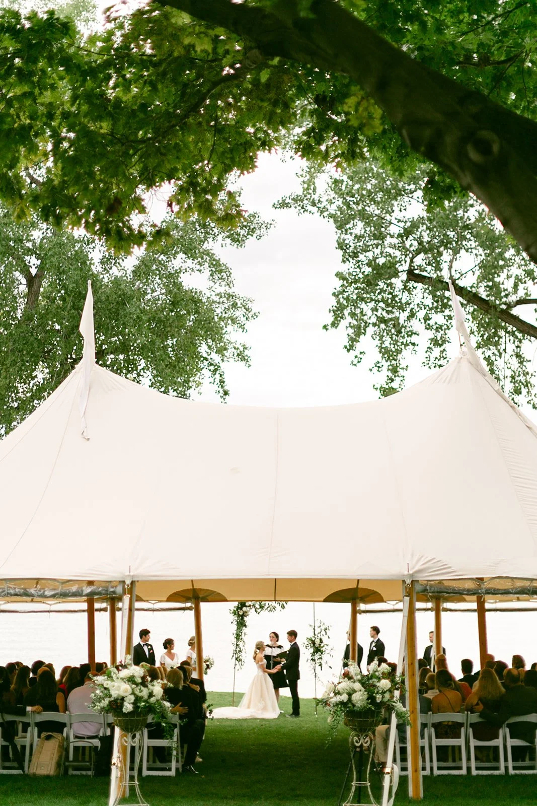 Outdoor wedding ceremony under a large white tent with guests seated, a couple at the altar, and trees in the background.