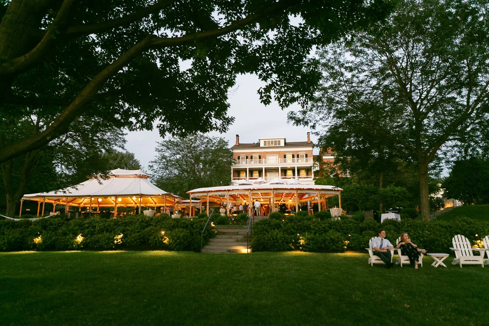 Large, well-lit house with a wraparound porch and outdoor seating, surrounded by trees and lawn, with people sitting in white chairs in the foreground.