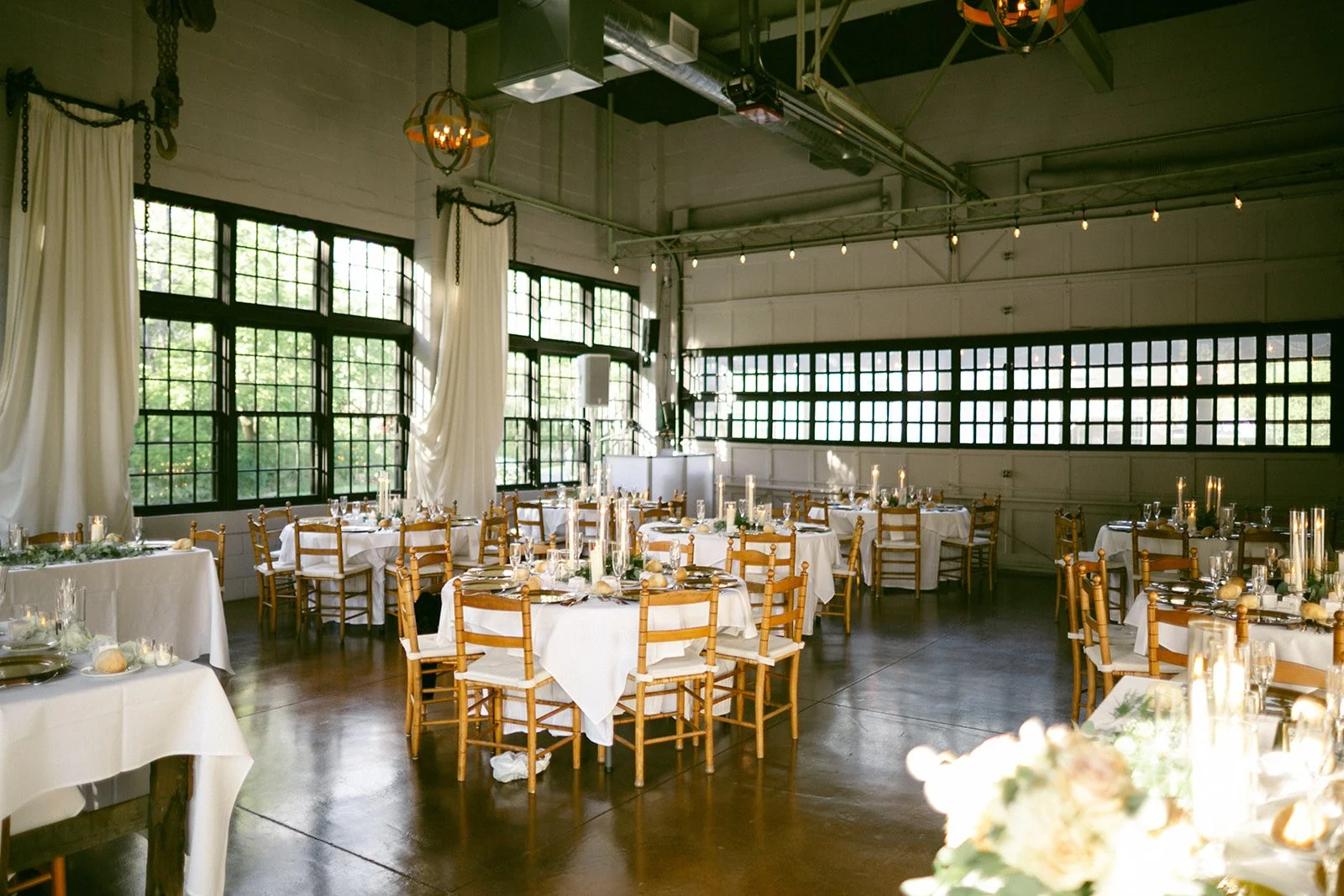 Sunlit dining room set for an event with round tables, wooden chairs, white tablecloths, candles, and large windows. Upstate New York Wedding Venue